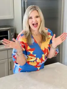 “Kelly Ettrich smiling and gesturing enthusiastically in a colorful dress while standing in a kitchen.”