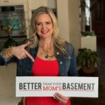 A smiling woman in a vibrant red dress and a denim jacket is standing in a well-lit kitchen. She's pointing to a sign she's holding, which reads "BETTER THAN YOUR MOM'S BASEMENT" in bold, contrasting fonts.