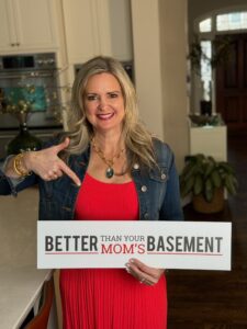 A smiling woman in a vibrant red dress and a denim jacket is standing in a well-lit kitchen. She's pointing to a sign she's holding, which reads "BETTER THAN YOUR MOM'S BASEMENT" in bold, contrasting fonts.