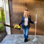 A professional woman with a warm smile, standing in front of a stylish wooden garage door, holding a leaf blower in one hand and a broom in the other, ready for spring cleaning. She is getting a home ready for the Loudoun County Real Estate Update!