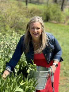"A woman in a red dress and a denim jacket smiles joyfully as she picks tulips at Hope Flower Farm and Winery. She is holding a gray bucket, ready to collect the flowers, with lush greenery and more tulip plants in the background under a clear blue sky."