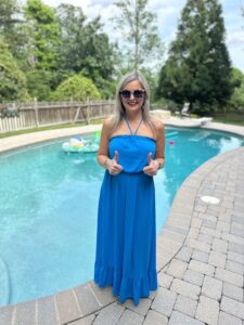 Kelly Ettrich standing by a pool, wearing a blue dress and sunglasses, giving two thumbs up and smiling.