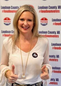 Kelly Ettrich giving a thumbs up in front of a Loudoun County voting backdrop with an 'I Voted' sticker on her shirt.