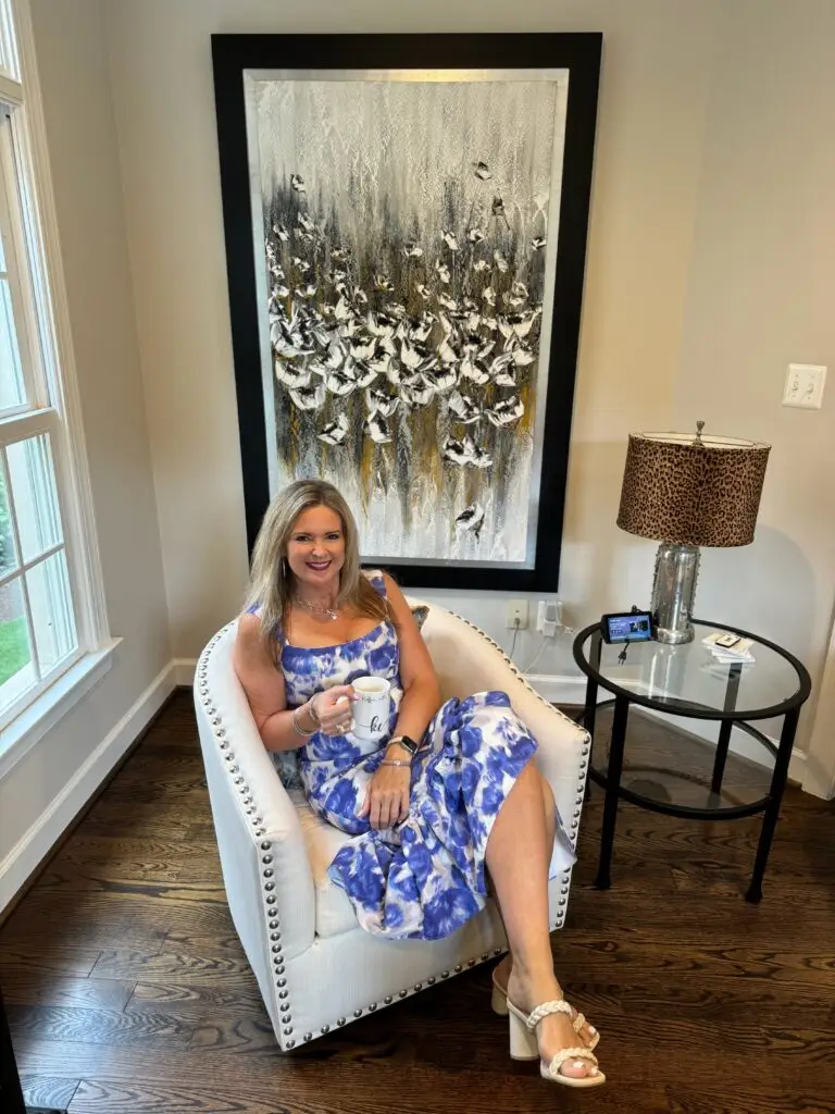 Kelly in a blue and white floral dress sitting on a white upholstered chair, holding a coffee mug, with a modern black-and-gold bird-themed painting in the background