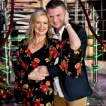 A smiling couple, Kelly and Mark, pose together on a grand staircase with glowing lights, dressed in elegant attire for a romantic Valentine’s Day celebration.