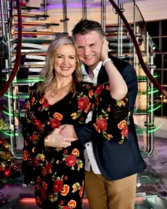 A smiling couple, Kelly and Mark, pose together on a grand staircase with glowing lights, dressed in elegant attire for a romantic Valentine’s Day celebration.