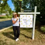 Kelly Ettrich Realtor Pointing to a For Sale Sign with A DayLight Savings Time Clock on the Cover